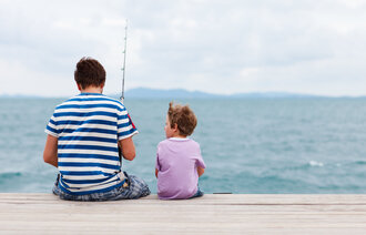 Et lite barn sitter sammen med en voksen mann på en brygge. Mannen fisker.