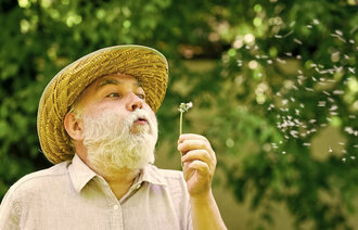 Bildet viser en eldre mann med stråhatt som blåser frøene av en løvetann