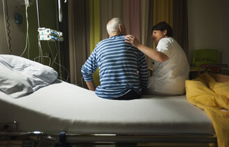 Bildet viser en sykepleier som sitter på sengekanten sammen med en eldre pasient