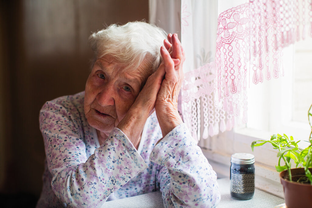 The photo shows an old, sad woman holding her head in her hands. On the table in front of her is a glass of pills.