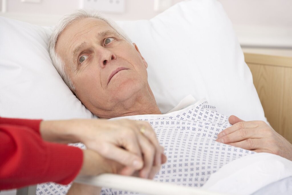Bildet viser en syk, eldre mann som ligger i en sykehusseng. En dames hender holder hans.