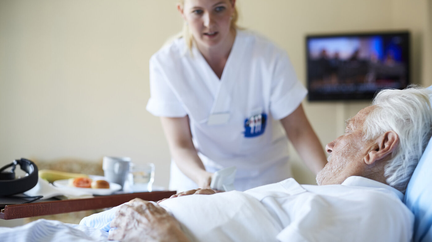 En sykepleier serverer en pasient mat på senga.