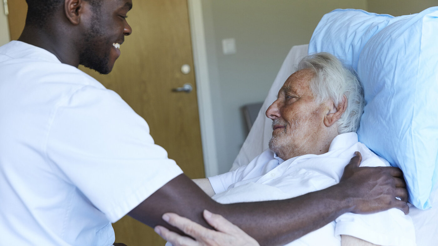 Bildet viser en mannlig sykepleier som smiler og holder hånden sin på skulderen til en liggende pasient