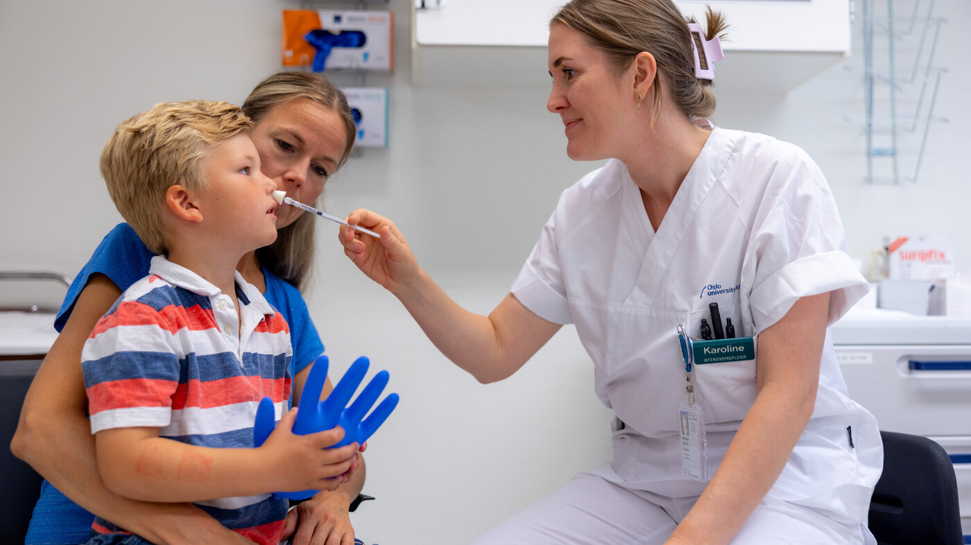 Nasal smertelindring til barn Bildet viser en liten gutt på fanget til sin mor. En sykepleier gir ham nesespray inn i det ene neseboret