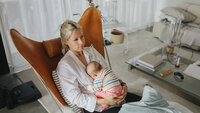 Bildet viser en nybakt mor som sitter i en lenestol hjemme. Hun ser litt oppgitt ut og har babyen liggende i fanget