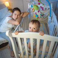 Bildet viser en mor som halvsover over barnesenga, mens barnet står i senga, lys våkent